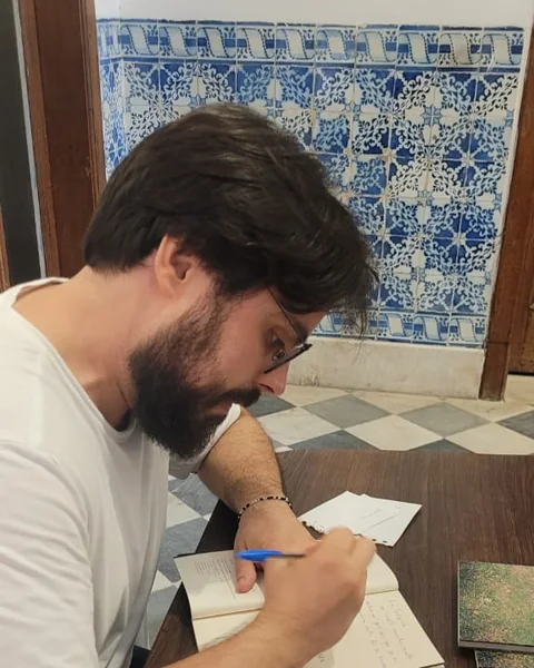 O autor, de camiseta branca, autografa um exemplar numa mesa de madeira, diante de uma parede de azulejos azuis e brancos.