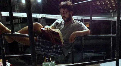Danilo de Athayde, de bermuda xadrez e camiseta cinza, lê um livro de capa vermelha sentado no alto de um andaime, à noite, com as pernas apoiadas numa das barras.