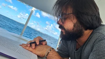 Danilo de Athayde, de perfil, lê um livro com uma caneta na mão, a bordo de um barco, com o mar e o céu azul ao fundo.