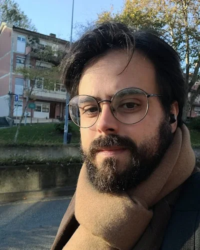 Danilo de Athayde, de óculos redondos e barba, com um cachecol caramelo, numa rua arborizada em dia de sol.